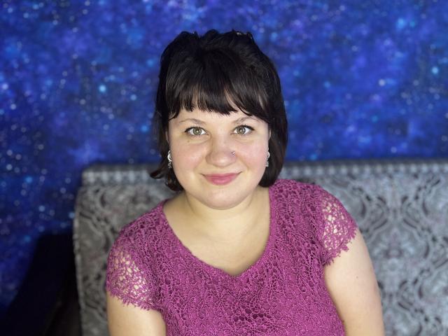 AlisaNAleks has short black hair, green eyes, a small nose ring, and a pink lace top, smiling slightly with her hands by her sides. She's a chubby chick.