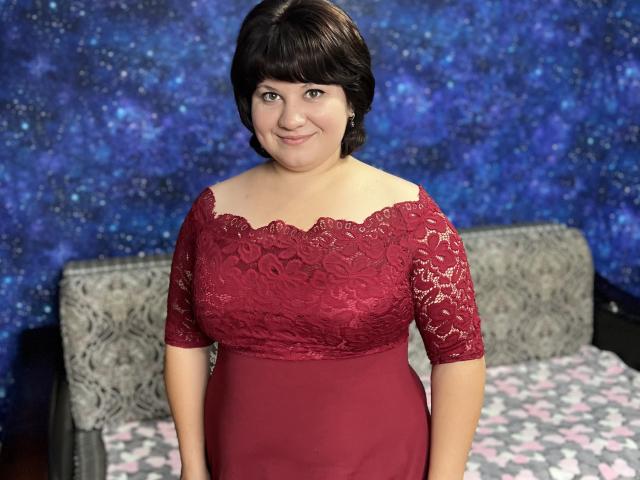 AlisaNAleks with short black hair, smiling, wearing a red lace-off shoulder dress, standing in front of a bed with gray pillows and a pink flower-patterned blanket.