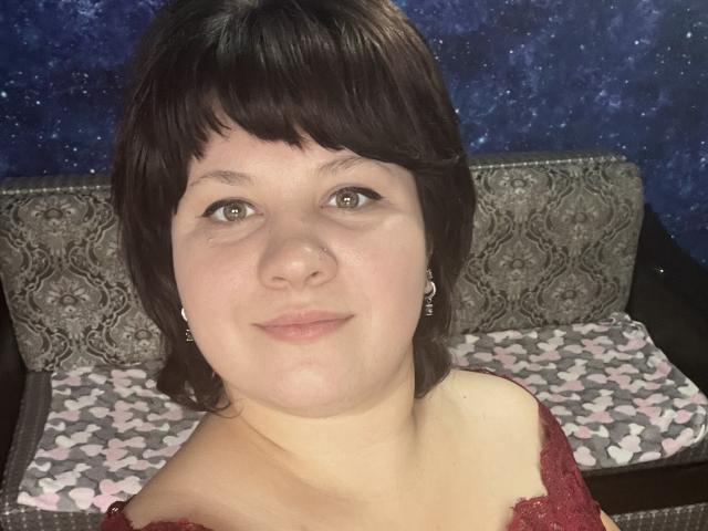 AlisaNAleks has short brown hair, green eyes, and a slight smile. She's wearing a red lace top with small earrings. Her tits are average-sized, and she's cute.