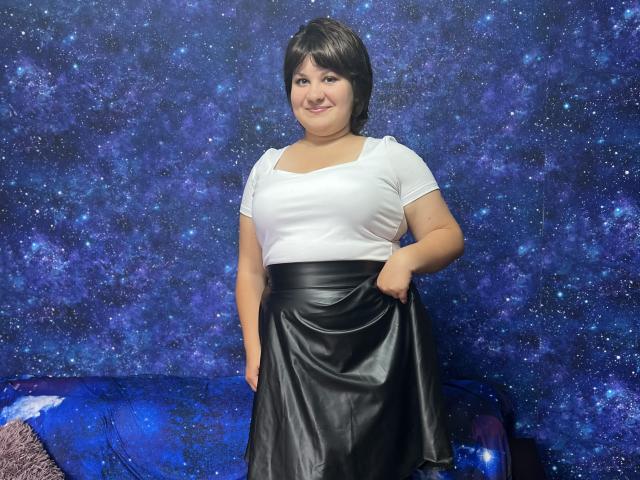 AlisaNAleks with short black hair, white tee, and black leather skirt, holding skirt, smiling, standing, blue starry background, and blue-patterned couch. Her tits are average-sized, curvy.