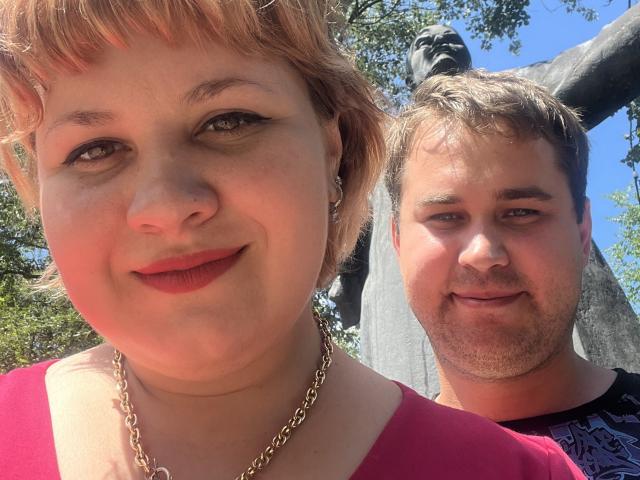 AlisaNAleks with short brown hair and red lipstick, wearing a pink top and gold chain, smiling at a guy with short brown hair and a black shirt.