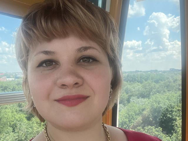 AlisaNAleks has short, brown hair, green eyes, and red lipstick. She's wearing a red top with a gold necklace. Her tits are small, and she's smiling slightly.