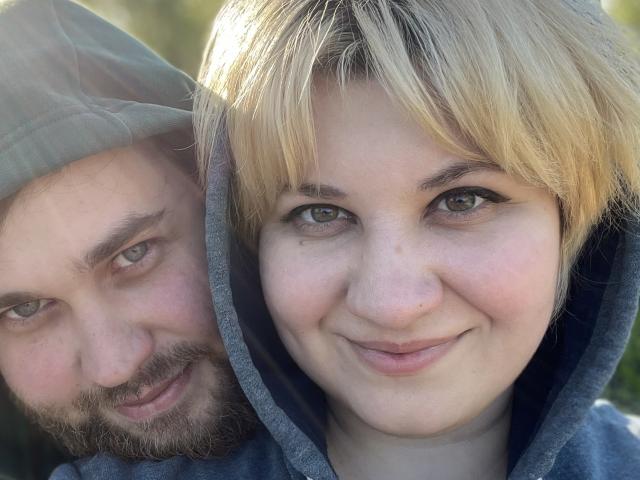 AlisaNAleks with blonde hair and green eyes, smiling, next to a bearded man in a green hat, both in grey hoodies. His face is partially in the background.