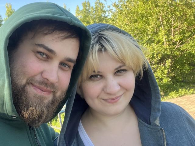 AlisaNAleks with a beard, green hoodie, and a bearded man, both smiling, standing close with blonde hair and blue hoodie, outdoors on a sunny day.