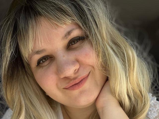 AlisaNAleks has blonde, wavy hair, fair skin, and is smiling softly with a slight nose bump. She's touching her neck with her right hand. Her eyes are green.