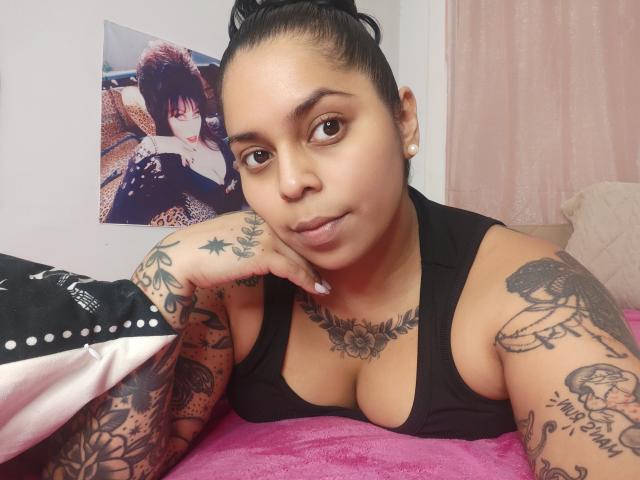 Chili está acostada sobre una manta rosa, apoyando la cabeza en la mano, con grandes ojos marrones mirando a la cámara. Lleva una camiseta sin mangas negra, mostrando sus tatuajes en los brazos y en el pecho, con el pelo recogido. Detrás de ella, un cartel de una mujer de aspecto similar es parcialmente visible.