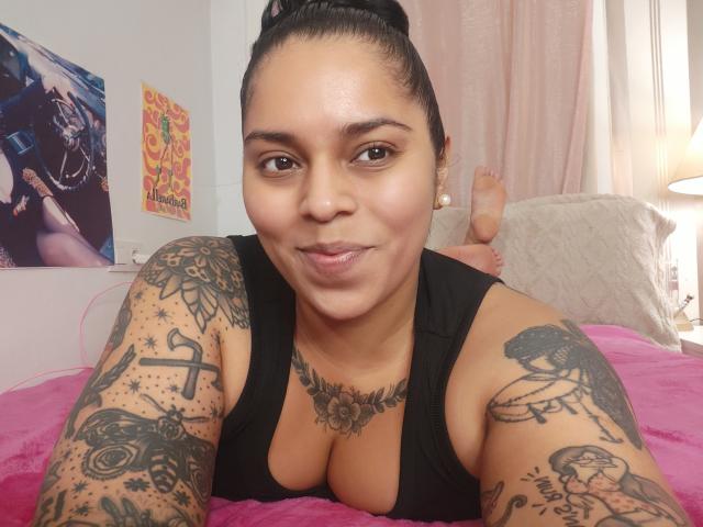 Chili, a curvy woman with dark skin and braided hair, lies on a bed with pink sheets, showing off her tattooed arms and chest, wearing a black tank top, smiling at the camera.