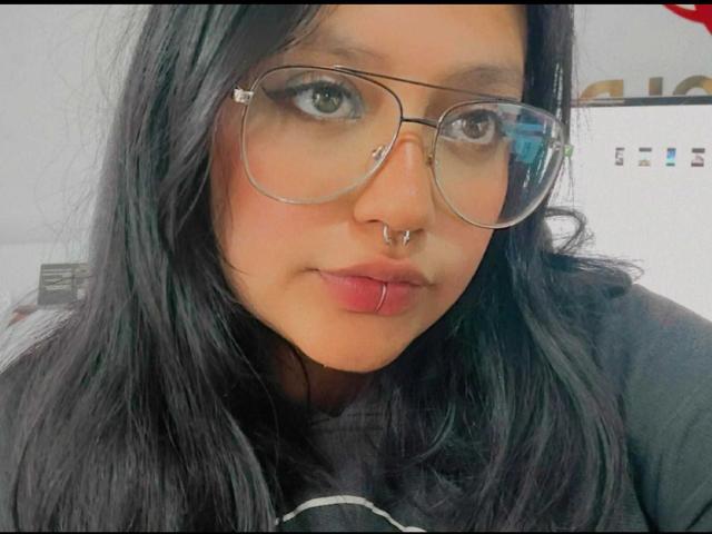 LilithAbrahel69 has black hair, glasses, a nose ring, and a lip ring. She's looking at the camera with a slight pout. She's wearing a black shirt.