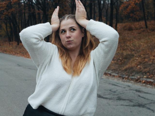 JessikaDaniels with hands on head, looking sideways, wearing a white knit sweater, black pants, and blonde hair, standing on a grey road, autumn leaves in background.