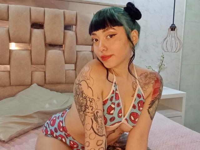 GigiVolkova with green-tipped hair in a bun, wearing a blue patterned bikini, showing tattoos on her arms, leaning on a bed with pink bedding, smiling.