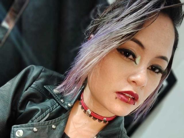 Cassie Jones, avec ses cheveux teints en violet, son maquillage vert, son rouge à lèvres rouge, sa lèvre inférieure percée, sa veste en cuir noir et son collier rouge, regarde l'appareil photo avec une légère inclinaison de la tête..