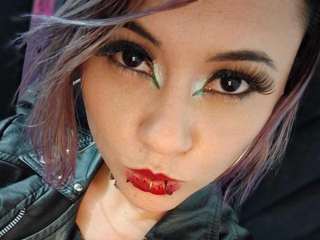 CassieJones has a goth face with green eyeshadow, purple hair, and a red lip piercing. She's wearing a black leather jacket and looking at the camera with pouty lips.