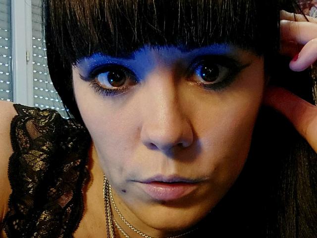 OrianaLaFrancaise has dark, straight bangs, blue eyeshadow, and a black lace top. She's wearing a chain necklace with a black mole under her lip. Her hand touches her hair.