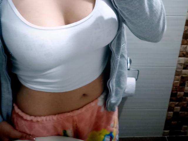 OdettePrincess in a white sports bra, showing her tits and a bit of her pink pajama bottoms, with a gray hoodie open and her hand on her hip.