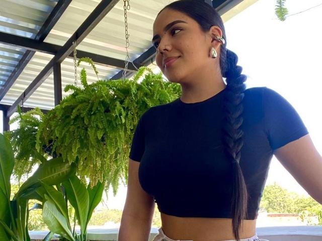 TamaraGray with long braided brown hair, black crop top, showing her tits, earrings, smiling sideways, hanging green plant, metal roof, sunny day, green plant leaves in background.