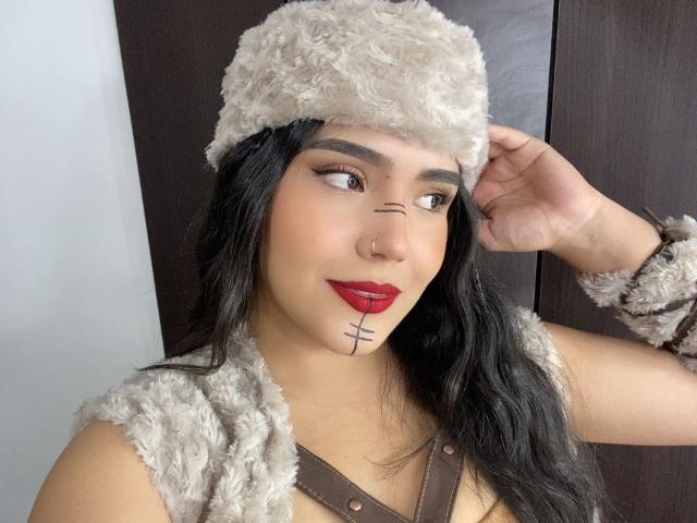 TamaraGray with long black hair, red lipstick, brown fur hat, and matching sleeve, has a black line tattoo under her right eye, wearing brown straps, touching her hat.