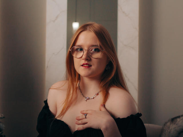 BonnieBell with red hair, glasses, and red lipstick, wearing a black off-shoulder top, holding her tits, necklace, ring, and a slight smile, in a soft-lit room.