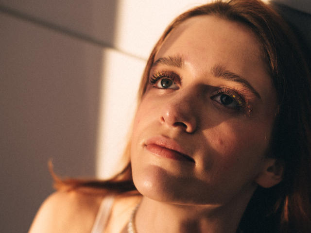 FrancesaL has fair skin, brown eyes, and red hair, with a slight blush on her cheeks, wearing a white tank top, gazing upwards with soft sunlight on her face.