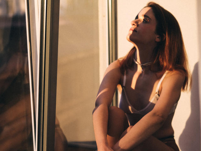 FrancescaL with red hair, wearing a white bra, sitting by a window, sunlight on her face, looking up, hands on knee, shadow on wall, subtle necklace.