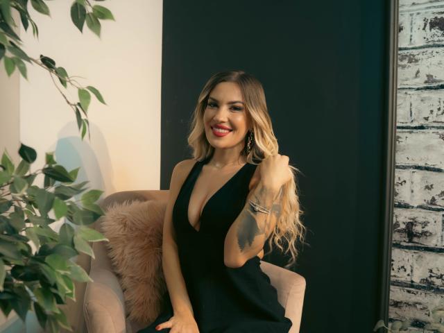 ZoeyMarie with long blonde hair, red lipstick, black deep cleavage dress, smiling, tattoo on left arm, sitting on pink chair with fluffy pillow, holding hair, green leafy plant.