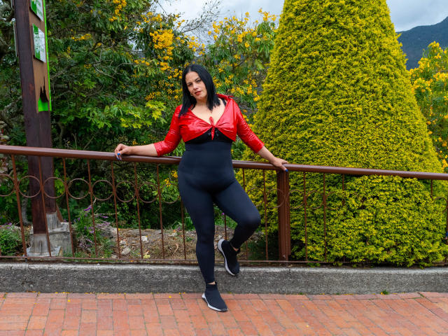 LizaRussell has black hair, red tied-up shirt showing her tits, black high-waisted pants, and black sneakers, leaning on a railing with a tall green bush behind her.