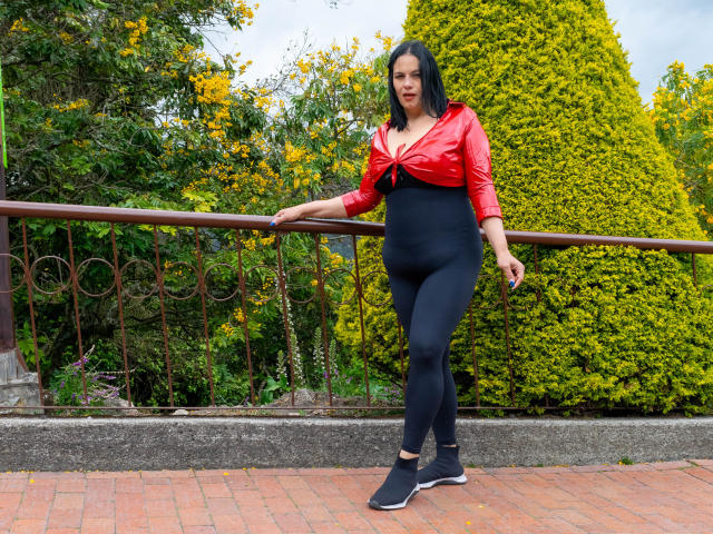 LizaRussell with medium tits, black high-waisted leggings, red tied-up top, black sneakers, leaning on brown railing, green and yellow bushy background, standing on red brick pavement.