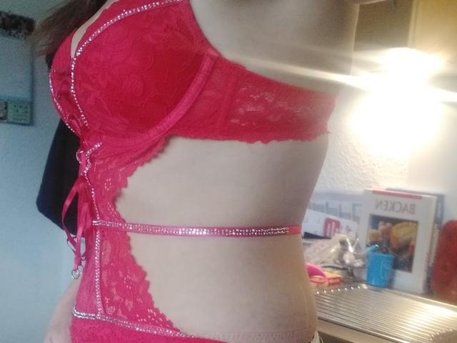 HotSexyLady69 in red lace lingerie with pink sequin accents, showing her side with visible tits and slim waist, in a brightly lit room.