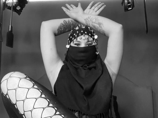 MalakCote, les bras croisés, portant un foulard orné de bijoux, un masque noir et un haut sans manches, laissant apparaître des tatouages, et un motif serpent sur la jambe, sur une photo en noir et blanc..