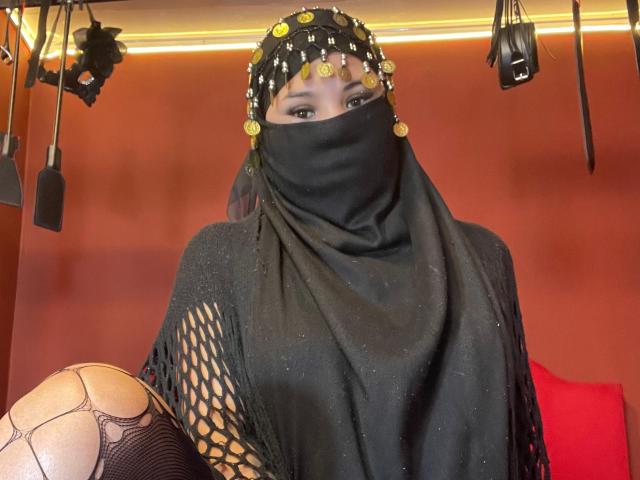 MalakCote with a black headscarf adorned with gold coins, black mask, and black fishnet shawl, showing a leg in fishnet stockings, in a BDSM-themed setting.