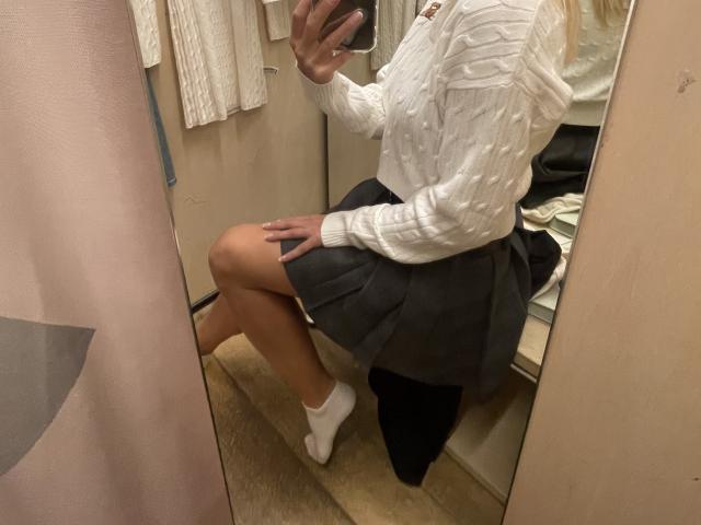 SandraSwweet sitting in a dressing room, wearing a white, textured, long-sleeve shirt and a grey, pleated skirt, with white socks, her ass slightly exposed.
