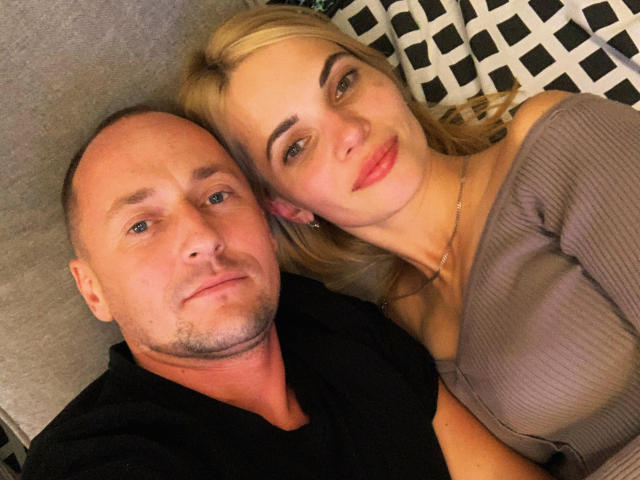 DonorDi (blonde woman) with her tits slightly visible, lying next to DonorDi (bald man) with a black shirt, both smiling, on a grey couch with a patterned pillow.