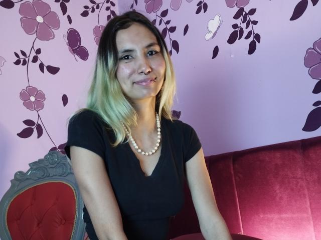 Ambeer69 has blonde-to-brown gradient hair, wearing a black top with a pearl necklace, sitting on a red velvet couch, smiling with a nose ring.
