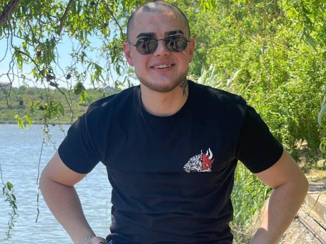 RichardSexy, bald with a tattoo on his neck, wearing black sunglasses and a tight black t-shirt with a red and white zebra logo, smiling by a river.