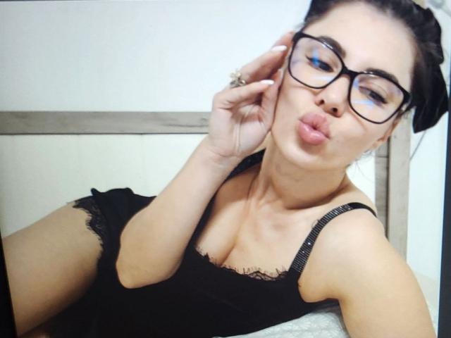Eilly with pouty lips, black glasses, and black lace top, showing off her small tits and cute face, hand in hair, sitting on bed, looking sexy.