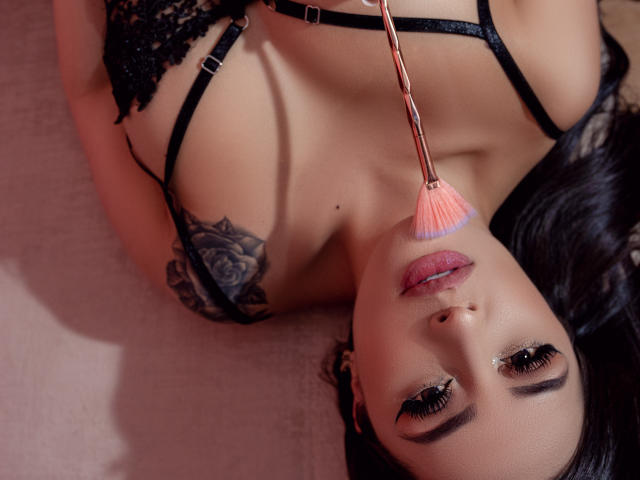 HannaMartinss with black lace bra, rose tattoo on shoulder, holding pink makeup brush to her pink-lipsticked, eyelined face, lying down, looking up. Her tits are visible.