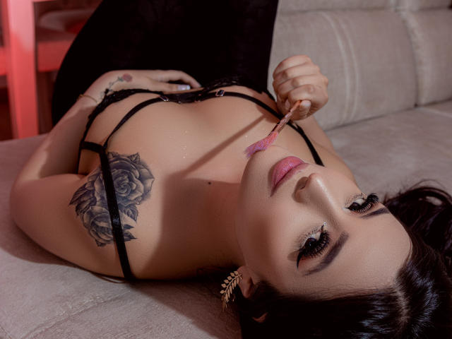 HannaMartinss lying on a couch, black lingerie, holding a pink lipstick to her slightly parted pink lips, black rose tattoo on her right shoulder, dark hair, subtle makeup.