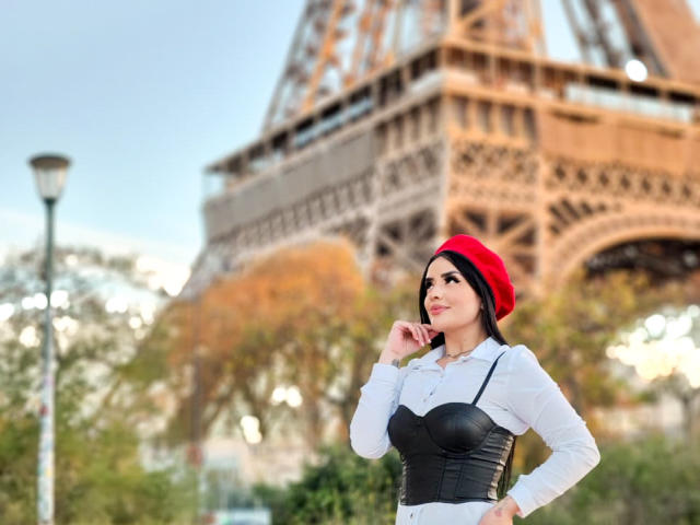 HannaMartinss with red beret, white shirt, black corset, hand on chin, looking up at Eiffel Tower, short black hair, cute, tits slightly visible, outdoors, daytime.