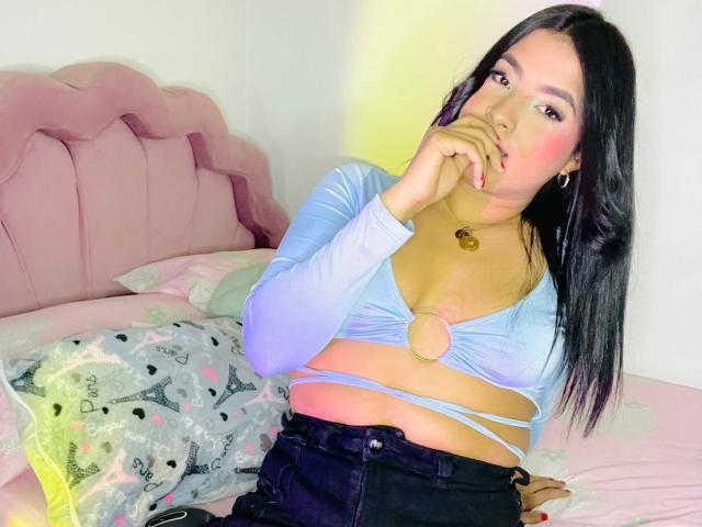 AbbieJoy with long black hair, wearing a blue top with a circular cutout, exposing her small tits, sits on a pink bed, finger near her pink-lipped mouth.
