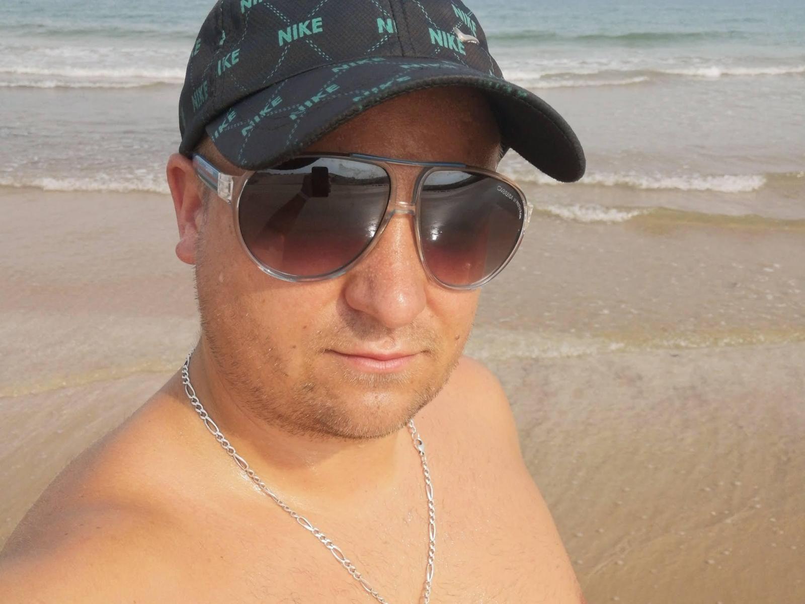 HornyJohnyO is shirtless, wearing a black Nike cap, sunglasses, and a silver chain, with a slightly tanned, hairy face and a neutral expression at the beach.