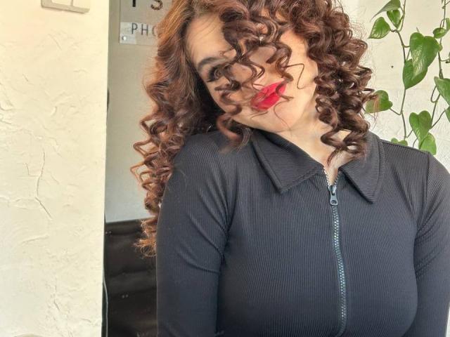 FridaCure with curly brown hair, red lipstick, and a black zip-up shirt, slightly tilting head with a playful smile, showing her big tits.