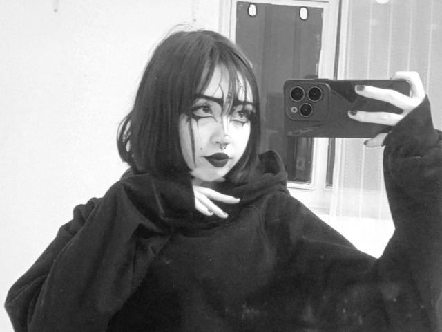 SilencePhsi, met donkere lipstick, zwarte oogmake-up en halflang zwart haar, maakt een selfie in een spiegel. Ze draagt een zwarte hoodie en houdt een telefoon met drie camera's vast..