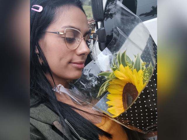 AnaisHilton with glasses, holding a sunflower bouquet, wearing a green jacket, black hair with pink hairclip, and a subtle smile. Her tits are partially visible under her jacket.