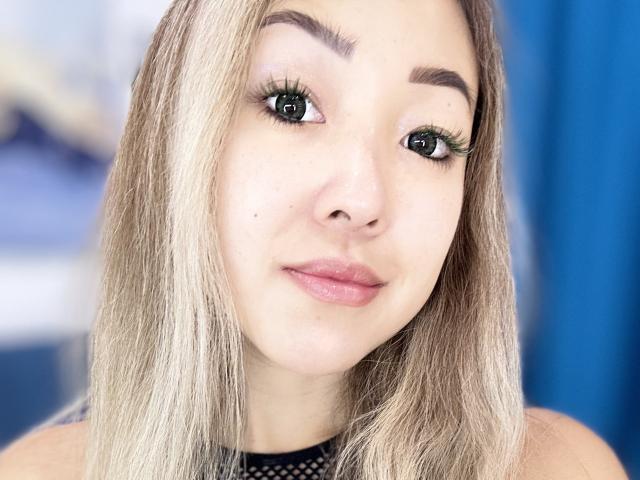 RenyLime has blonde hair, green eyes with long eyelashes, pink lips, and a small mole on her left cheek. She's wearing a black mesh top, looking at the camera.