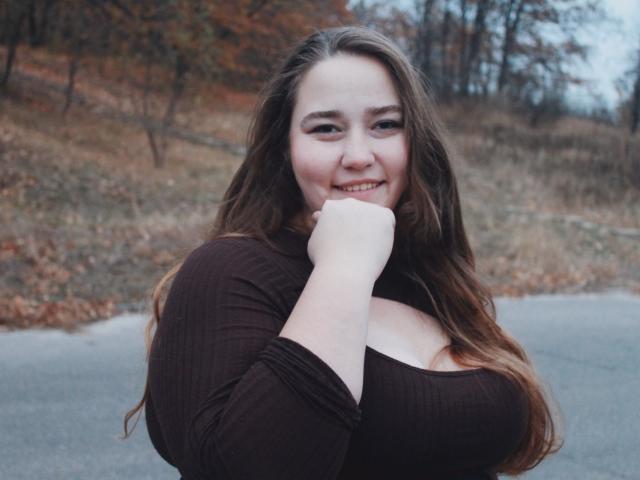 DaniellaGraceZZ, with big boobs, smiling, brown hair, wearing a ribbed brown top, hand near her mouth, standing outside, overcast day, soft light, gentle expression.
