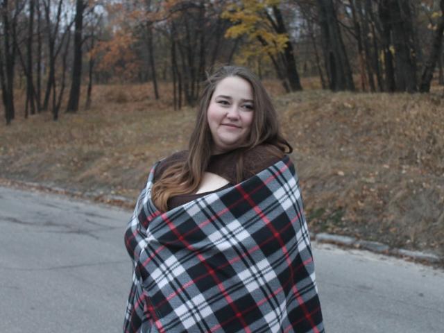 DaniellaGraceZZ with long brown hair, smiling, wrapped in a black, white, and red plaid blanket, brown shirt underneath, standing on a grey, wet sidewalk.
