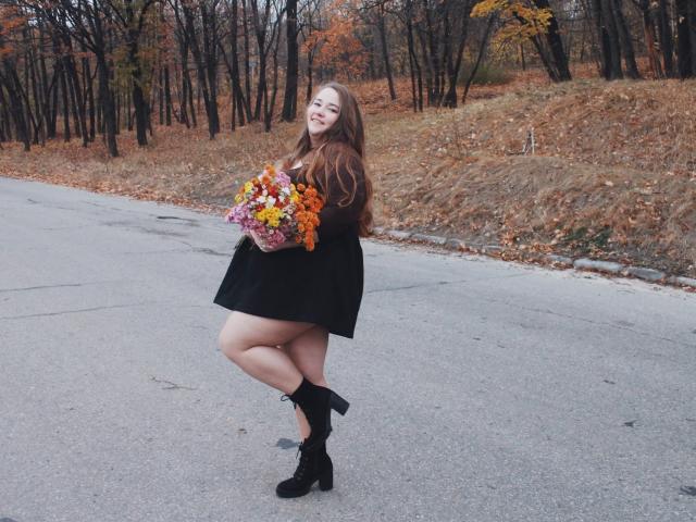 DaniellaGraceZZ, with long brown hair, black dress, and black boots, holding a colorful bouquet, stands on one leg, smiling, showing her curvy ass and fluffy tits.