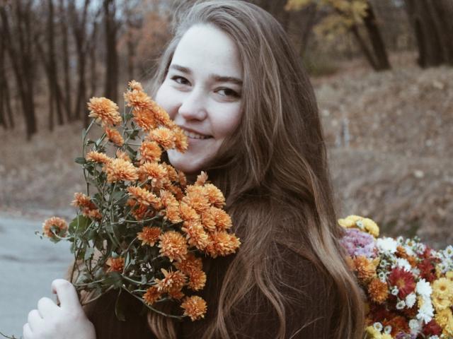 DaniellaGraceZZ with long brown hair, holding a bouquet of orange chrysanthemums, smiling, wearing a brown sweater, autumn forest in the background, soft natural light.