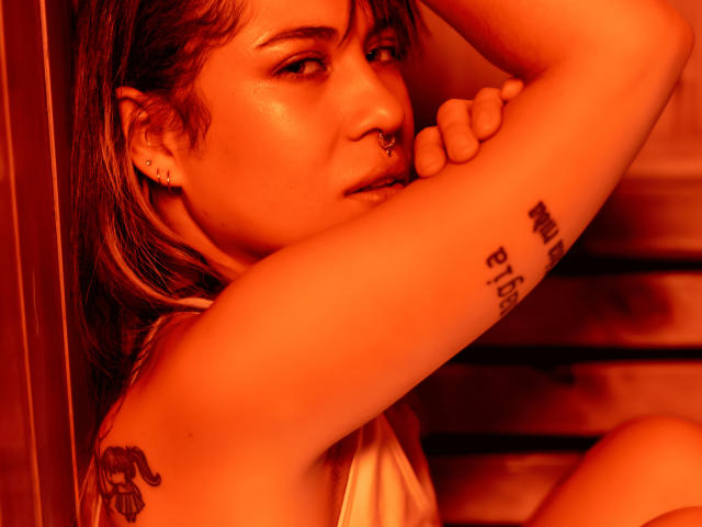 HellenK with a nose ring, ear piercings, and tattoos, in a warm orange light, looking over her shoulder with her arm raised, showing her armpit tattoo.