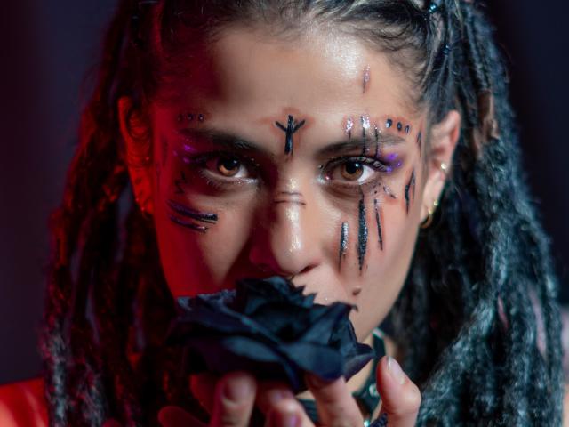 HellenK, avec ses cheveux noirs et bouclés, tenant un tissu noir, a des rayures et des points de maquillage noirs sur son visage, un éclairage rouge et violet, et une expression sérieuse..
