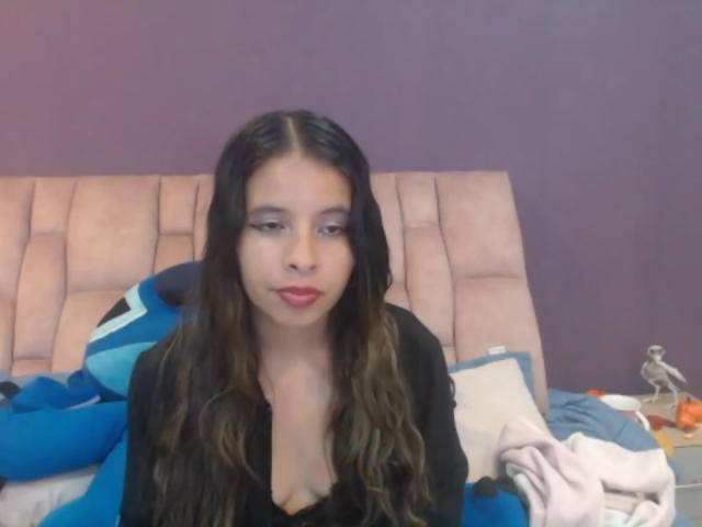 JuliaWaine with long, wavy brown hair, red lipstick, and a black top, sitting on a bed with blue and white pillows, and a pink blanket. Her tits are small.